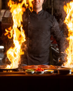 Chef con chaqueta negra cocinando con dos sartenes que producen grandes llamaradas, frente a una barra con ingredientes frescos como tomates cherry, vegetales y salsas.