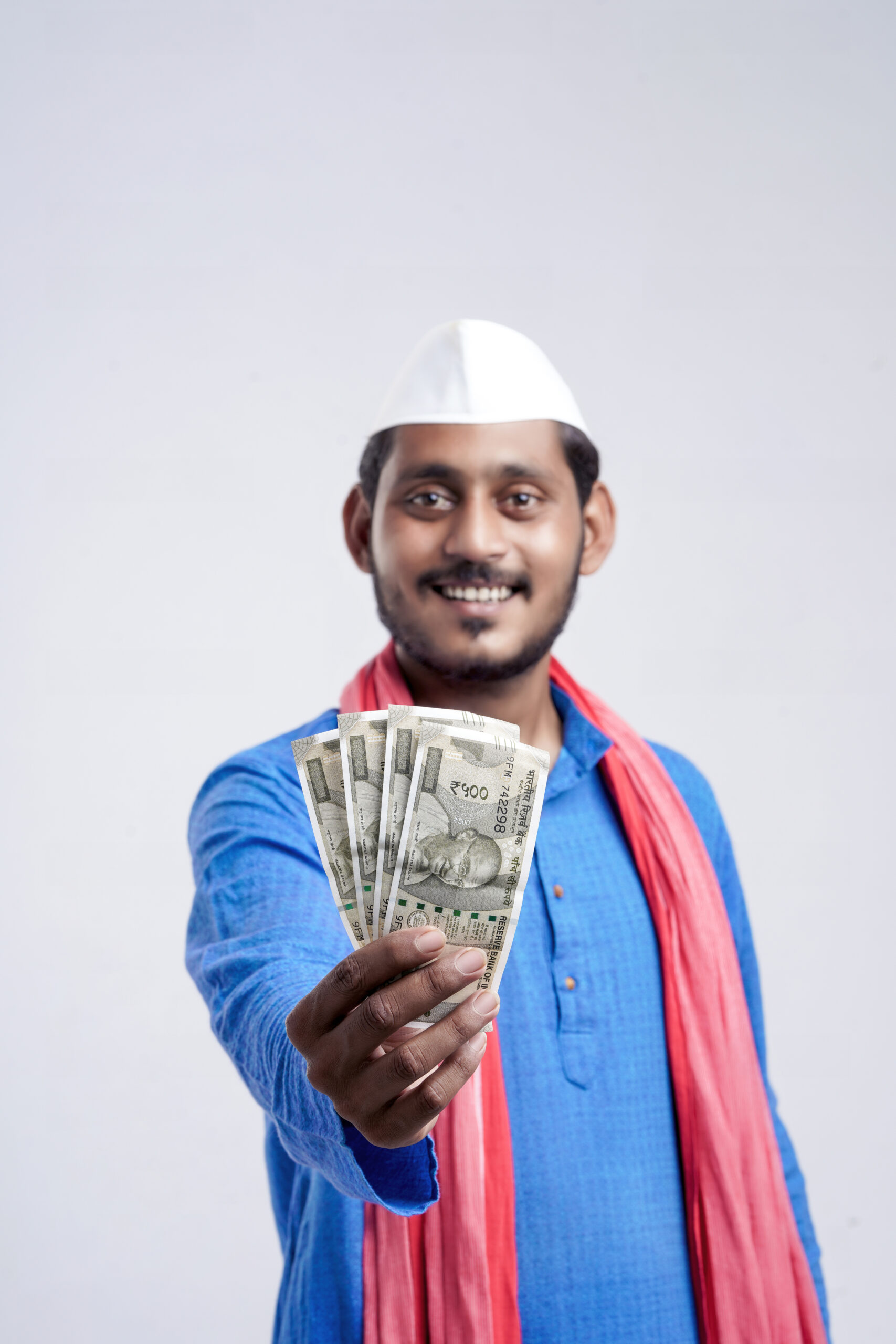 Hombre joven indio con gorro blanco y bufanda roja, sosteniendo billetes de 500 rupias hacia la cámara.