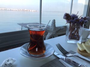 Vaso de té turco sobre una mesa blanca, junto a un plato con queso, flores secas en un florero y el mar visible a través de una ventana.